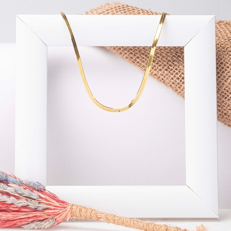 May include: A gold chain necklace displayed in a white frame. The chain is a flat, thin, and shiny design.