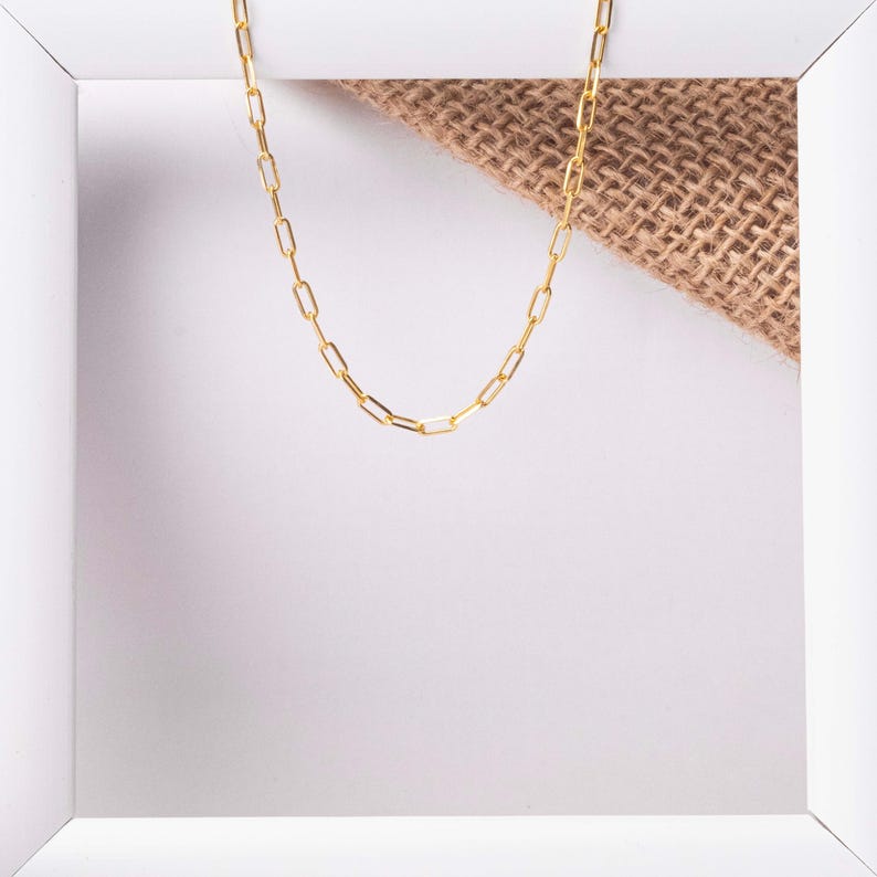 May include: A gold chain necklace with a simple, linked design. The chain is displayed on a white background with a piece of burlap fabric.
