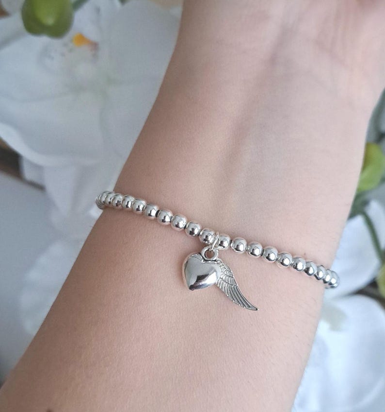 May include: A silver beaded bracelet with a heart and wing charm. The bracelet is made of small, round, silver beads. The charm is a heart with a single wing attached. The bracelet is worn on a wrist.