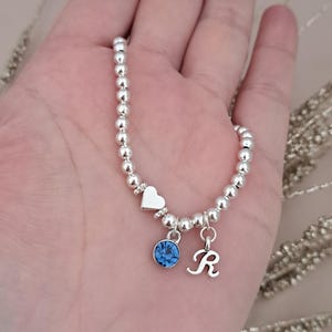 Może przedstawiać: Srebrna bransoletka z koralik&oacute;w z zawieszką w kształcie serca, niebieskim kamieniem szlachetnym i zawieszką z literą "R". Bransoletka jest trzymana w dłoni, ukazując delikatny design i połysk metalu.