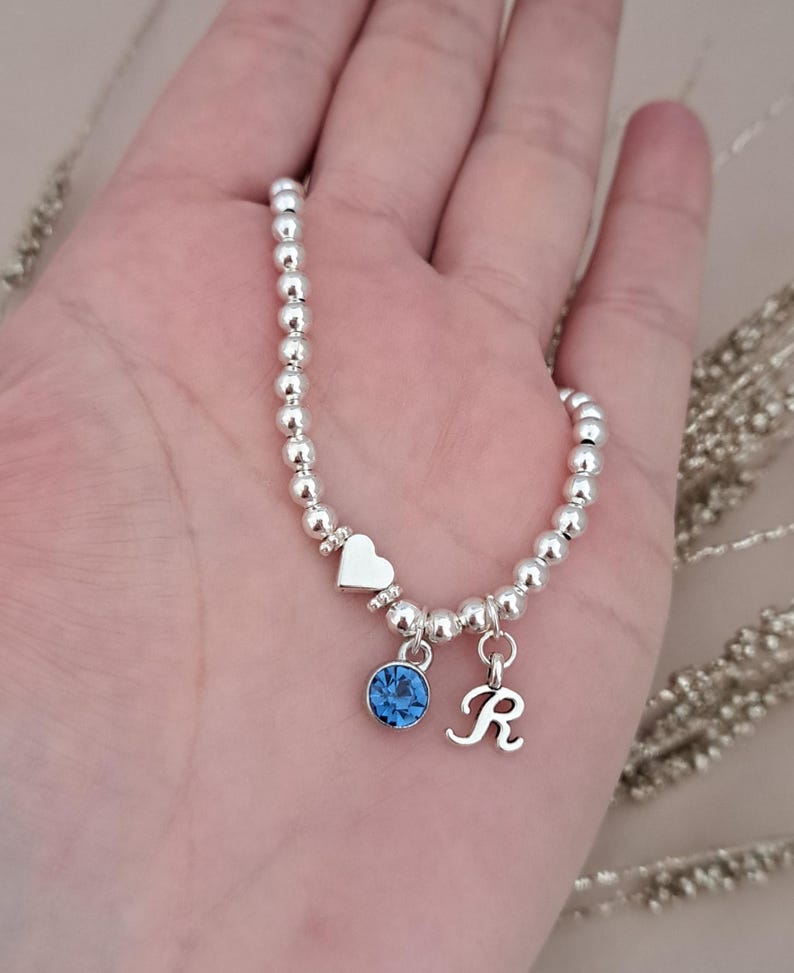 Może przedstawiać: Srebrna bransoletka z koralik&oacute;w z zawieszką w kształcie serca, niebieskim kamieniem szlachetnym i zawieszką z literą "R". Bransoletka jest trzymana w dłoni, ukazując delikatny design i połysk metalu.