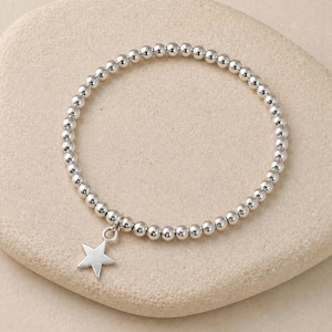 May include: Silver beaded bracelet with a star charm. The bracelet is made of small, round silver beads, with a five-pointed star pendant hanging from the centre. The bracelet is shown on a light-coloured, textured surface.