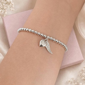 May include: Silver beaded bracelet with a heart and wing charm. The bracelet features small, round beads and a dangling charm with a polished heart and a detailed wing. The bracelet is displayed on a light pink surface.