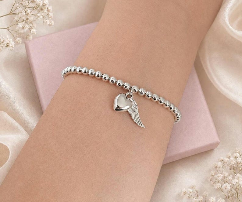 May include: Silver beaded bracelet with a heart and wing charm. The bracelet features small, round beads and a dangling charm with a polished heart and a detailed wing. The bracelet is displayed on a light pink surface.