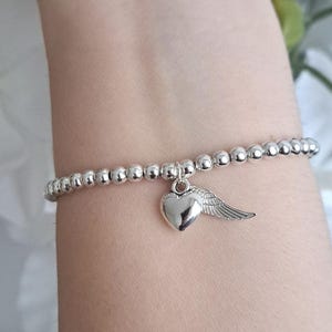May include: Silver beaded bracelet with a heart and wing charm. The bracelet features small, round silver beads strung together. A heart-shaped charm with a wing detail hangs from the bracelet, adding a touch of elegance. The bracelet is shown on a wrist.