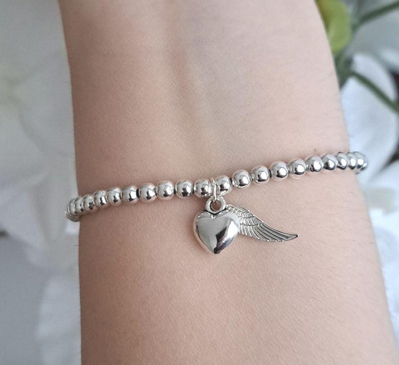 May include: Silver beaded bracelet with a heart and wing charm. The bracelet features small, round silver beads strung together. A heart-shaped charm with a wing detail hangs from the bracelet, adding a touch of elegance. The bracelet is shown on a wrist.