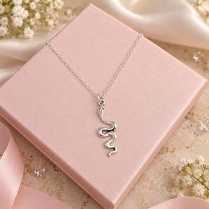 May include: A silver-coloured metal necklace featuring a snake-shaped pendant. The pendant is suspended from a delicate, thin silver-coloured chain. The snake design is smooth and elongated, with a defined head and a curved, winding body shape.