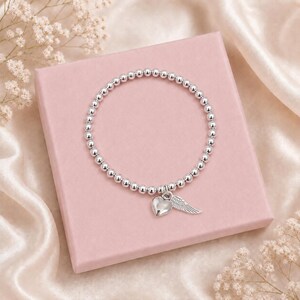 May include: Silver beaded bracelet with a heart and wing charm. The bracelet is displayed on a pink box, with a satin background and small white flowers.