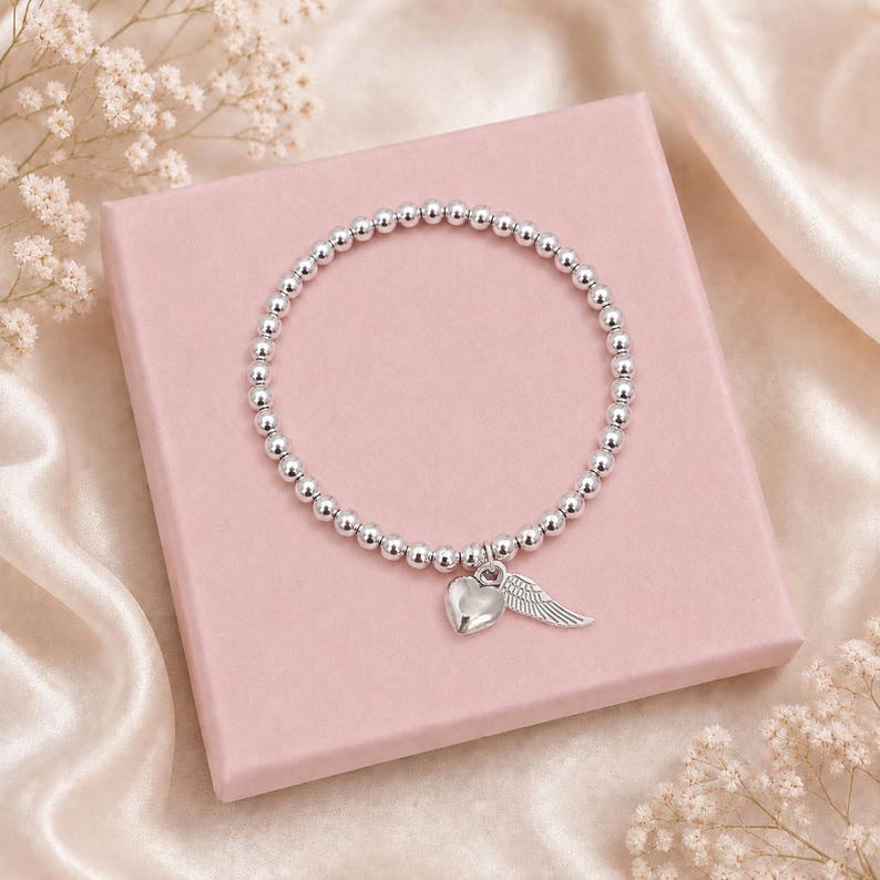 May include: Silver beaded bracelet with a heart and wing charm. The bracelet is displayed on a pink box, with a satin background and small white flowers.
