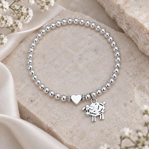 May include: A silver-toned beaded bracelet featuring a small, smooth heart-shaped charm and a dangling sheep-shaped pendant. The bracelet is constructed from a continuous strand of uniform, polished metallic spheres arranged in a circular loop.