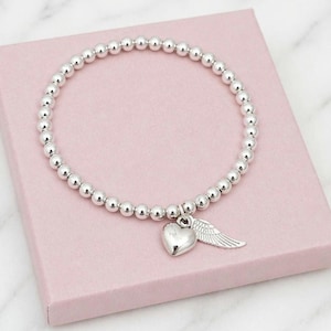 May include: Silver beaded bracelet with a heart and wing charm. The bracelet features small, round silver beads strung together. A silver heart charm and a wing charm dangle from the bracelet. The bracelet is displayed on a pink box.