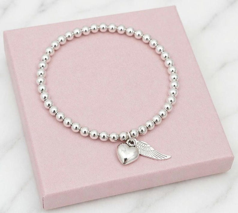 May include: Silver beaded bracelet with a heart and wing charm. The bracelet features small, round silver beads strung together. A silver heart charm and a wing charm dangle from the bracelet. The bracelet is displayed on a pink box.