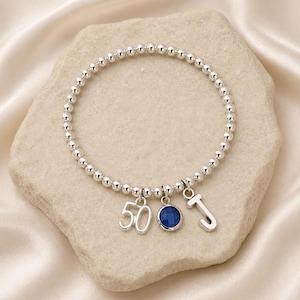Op de afbeelding: Zilveren kralenarmband met een blauwe edelsteenbedel, de bedel met het nummer "50" en een letter "J" bedel. De armband wordt gepresenteerd op een lichtgekleurd stenen oppervlak.