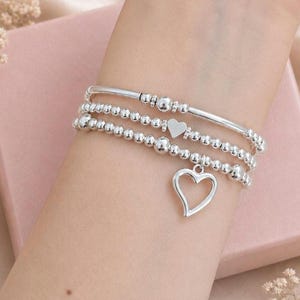 May include: Three silver bracelets are stacked on a wrist. The top bracelet is a simple bar, the middle has a small heart, and the bottom has a heart charm. The bracelets are on a wrist with a pink box in the background.