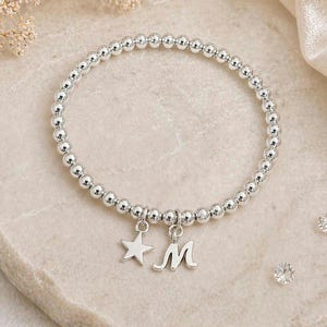 Op de afbeelding: Zilveren kralenarmband met een sterbedel en een letter "M" bedel. De armband is gemaakt van kleine, ronde, zilveren kralen. De bedels zijn ook zilverkleurig en hangen aan de armband.