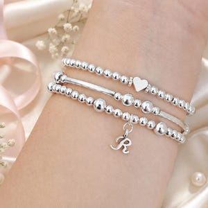 May include: A set of three silver-toned beaded bracelets. The top bracelet features a small heart charm. The middle bracelet includes a smooth cylindrical bar. The bottom bracelet features a dangling letter R charm. All beads are metallic and round.