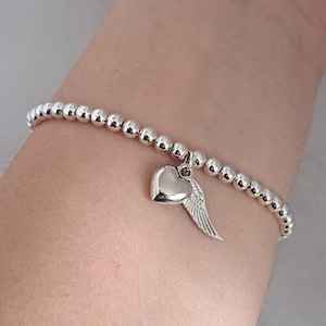 May include: Silver beaded bracelet with a heart and wing charm. The bracelet features small, round beads and a polished heart pendant with a detailed wing design. The bracelet is worn on a wrist.
