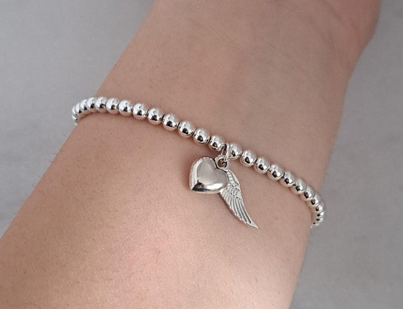 May include: Silver beaded bracelet with a heart and wing charm. The bracelet features small, round beads and a polished heart pendant with a detailed wing design. The bracelet is worn on a wrist.