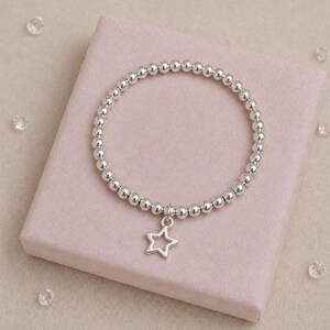 May include: Silver beaded bracelet with a star charm. The bracelet is made of small, round silver beads, with a hollow star pendant. The bracelet is presented on a pink surface.