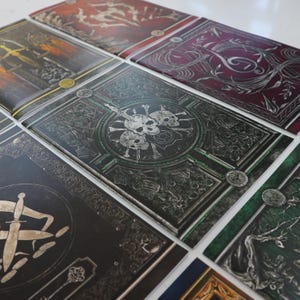 May include: A selection of fantasy-themed journals with detailed, embossed covers. The covers display skulls, swords, and gothic designs in silver, black, green, and red. The journals are arranged in a grid formation.