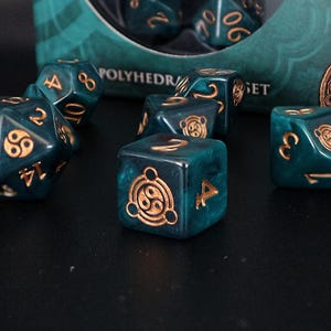 Op de afbeelding: Een set polyhedrale dobbelstenen in een teal en goud kleurenschema. De dobbelstenen hebben goudkleurige cijfers en symbolen. De dobbelstenen zijn gerangschikt op een donkere ondergrond. De doos op de achtergrond leest "POLYHEDR" en "SET".
