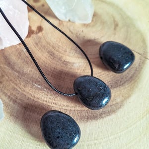 Könnte beinhalten: Eine schwarze Steinanhänger-Halskette mit einem passenden Stein und zwei zusätzlichen schwarzen Steinen. Die Halskette ist an einem schwarzen Band. Die Steine liegen auf einer Holzoberfläche mit Kristallen im Hintergrund.