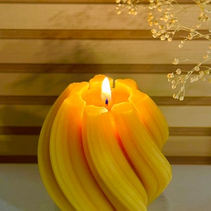 Puede incluir: Una vela amarilla en espiral encendida. La vela tiene un diseño texturizado y acanalado con una llama brillante. El fondo presenta una superficie clara y rayada y una ramita de flores secas. La vela es un artículo decorativo.