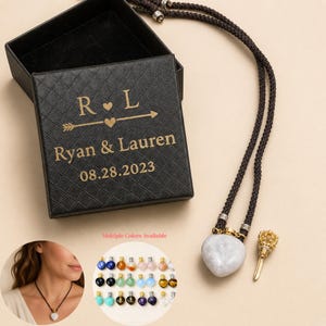 Puede incluir: Una caja de regalo cuadrada negra con textura acolchada, con texto dorado que dice R L, Ryan & Lauren y 08.28.2023. Al lado hay un colgante de piedra en forma de corazón en un cordón marrón oscuro, con un tapón dorado en forma de corona.