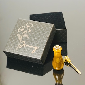Könnte beinhalten: Eine offene schwarze Geschenkbox mit einem grauen Deckel mit floralem Design und dem Namen "Jenny" in goldener Schrift. Ein kleiner, bernsteinfarbener Flaschenverschluss und ein goldfarbener Schlüssel liegen neben der Box.