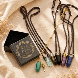 May include: A black gift box with gold text reading "Alice & David 08.15.2021" is open, next to several crystal pendant necklaces with brown cords. The pendants are various colours, including green, clear, black, purple, and blue.
