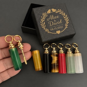 May include: A set of cylindrical stone vials with gold-toned metal caps and attached rings, including green, yellow, black, tiger eye, red, light green, and white stones. A black gift box features a gold laurel wreath and the text Alice & David 08.15.2021.