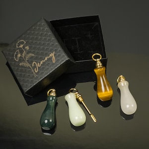 May include: An open black gift box with the name "Jenny" in gold script and floral accents. Four gemstone pendants with gold-colored metal tops are displayed. The pendants are green, white, yellow, and a tiger's eye pattern.