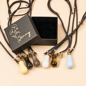May include: A black jewellery box with the gold text Jenny and a floral illustration, accompanied by several teardrop-shaped pendants in various colours including white, black, grey, tiger eye, and light blue, all attached to dark brown braided cords.