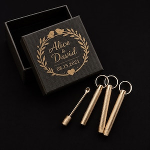 May include: A black square gift box with a textured diamond pattern and gold foil engraving reading Alice & David 08.15.2021 inside a bird and leaf wreath. Three gold-toned metal snuff spoons with knurled handles and ring tops are placed nearby.