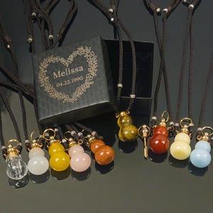 May include: A collection of gourd-shaped gemstone pendants on black cords. The pendants are in various colors, including clear, white, yellow, pink, and orange. A black box with the name "Melissa" and the date "04.22.1990" is also present.