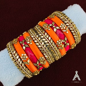 May include: A set of orange and pink silk thread bangles with gold and silver embellishments. The bangles are decorated with floral and leaf designs and have clear rhinestones.