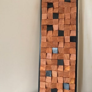 Op de afbeelding: Een rechthoekig wandkunstwerk gemaakt van hout. Het hout is gerangschikt in een rasterpatroon, met enkele vierkanten die zwart zijn geverfd. Het stuk is omlijst in zwart.