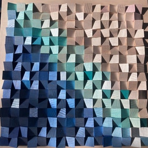 Op de afbeelding: Een wandkunstwerk gemaakt van hout met een gradiënt van blauwe, groene en beige kleuren. Het hout is gerangschikt in een geometrisch patroon, waardoor een textuur en een moderne uitstraling ontstaat.