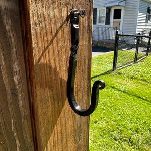 May include: A black metal hook is screwed into a wooden post. The hook is twisted and has a rounded end.