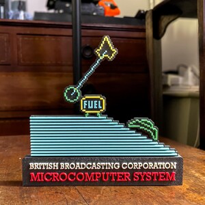 May include: A black and red 3D printed model of a retro video game. The model features a blue and yellow spaceship with a fuel tank labelled "FUEL". The model is on a black base with the text "BRITISH BROADCASTING CORPORATION MICROCOMPUTER SYSTEM".