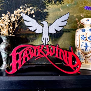 May include: A red and black wooden sign featuring the word "Hawkwind" in a stylized font, with a silver dove above. The sign is displayed on a black surface.