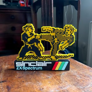Könnte beinhalten: Eine gelbe und schwarze 3D-gedruckte Figur von zwei Figuren in Kampfpose. Die Basis der Figur ist schwarz mit dem Text "Sinclair ZX Spectrum" in weiß.