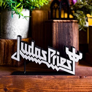 May include: A black and silver 3D printed sign featuring the text Judas Priest in a stylized font. The letters have a textured silver front surface with black edges and are mounted on a thin black rectangular base stand for display purposes.