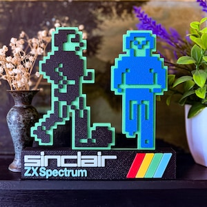 Könnte beinhalten: Retro-Schreibtischdekoration mit zwei pixeligen Figuren in Schwarz und Blau vor grünem Hintergrund. Der Sockel zeigt den Text "sinclair ZX Spectrum" und ein farbenfrohes Streifendesign. Ein nostalgisches Stück für Gamer.