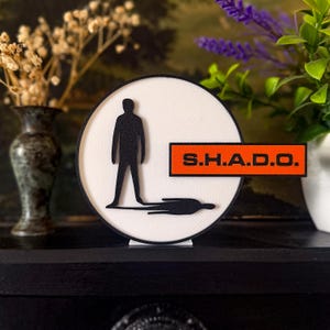 Può includere: Un oggetto decorativo rotondo, bianco e nero, con una silhouette nera di una persona e il testo "S.H.A.D.O." su un rettangolo arancione. Il design include un effetto ombra. L'oggetto è esposto su una superficie nera.