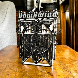 Könnte beinhalten: Eine schwarz-weiße 3D-gedruckte Skulptur des Bandlogos von Hawkwind. Das Logo zeigt einen stilisierten Adler mit ausgebreiteten Flügeln und einen Kreis mit Sternen drumherum. Die Skulptur steht auf einer Holzoberfläche.