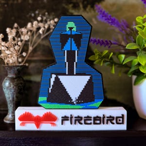 Peut inclure: Une œuvre d'art pixelisée représentant une figure bleue et noire, un triangle blanc et une tête verte. La base porte l'inscription "FIREBIRD" en noir avec un emblème d'oiseau rouge. L'œuvre est présentée sur un socle blanc.