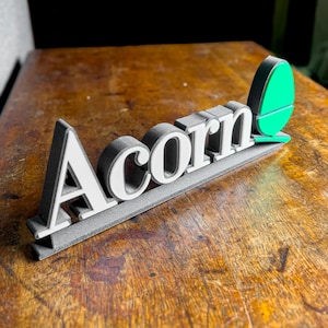 May include: A grey and white sign with the word "Acorn" in white letters. A green acorn is on the right side of the sign.