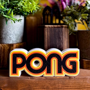 Può includere: Un'insegna tridimensionale che presenta la parola PONG in lettere nere in grassetto. Il lettering è delineato con strati di arancione brillante e giallo, creando un effetto a strati. L'insegna ha una base bianca e una finitura superficiale testurizzata.