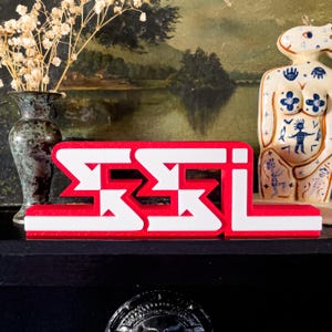 Op de afbeelding: Een rood en wit decoratief bord met de letters "SSL" in een vet, gestileerd lettertype. Het bord staat op een zwart oppervlak, met een vintage vaas en een decoratief beeldje op de achtergrond.